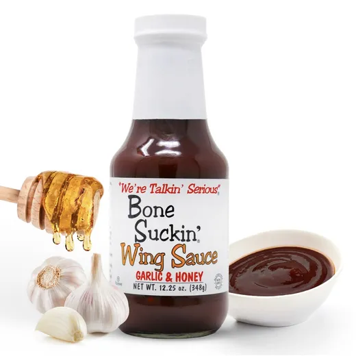 A bottle of Bone Suckin' Wing Sauce Garlic & Honey beside garlic bulbs, honey, and a small bowl of sauce.